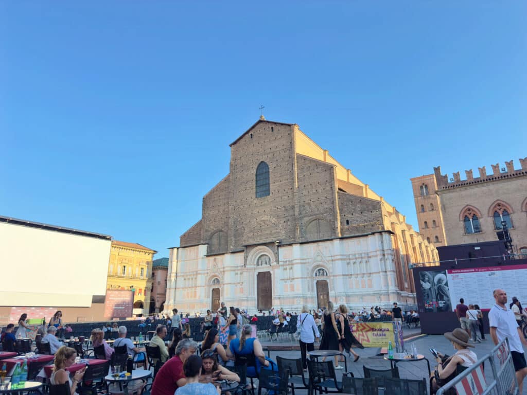 People sit at outdoor tables in a busy square with the Basilica of San Petronio in Bologna, Italy, in the background under a clear blue sky. Other historic buildings and event signage are also visible.