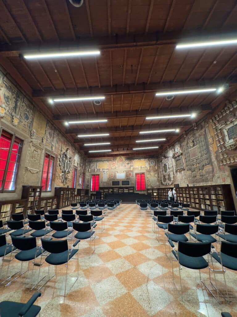 A large, ornate library room, Stabat Mater Hall of the Archiginnasio, with high wooden ceilings, decorated walls, red curtained windows, rows of black chairs, bookshelves lining the walls, and a checkerboard tile floor. One person stands near the bookshelves.