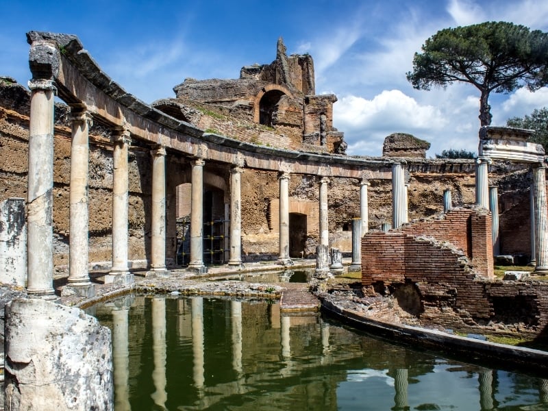 Ancient Roman ruins with tall stone columns and weathered brick walls, surrounding a reflective pool of water, set against a blue sky with clouds and a large pine tree in the background.