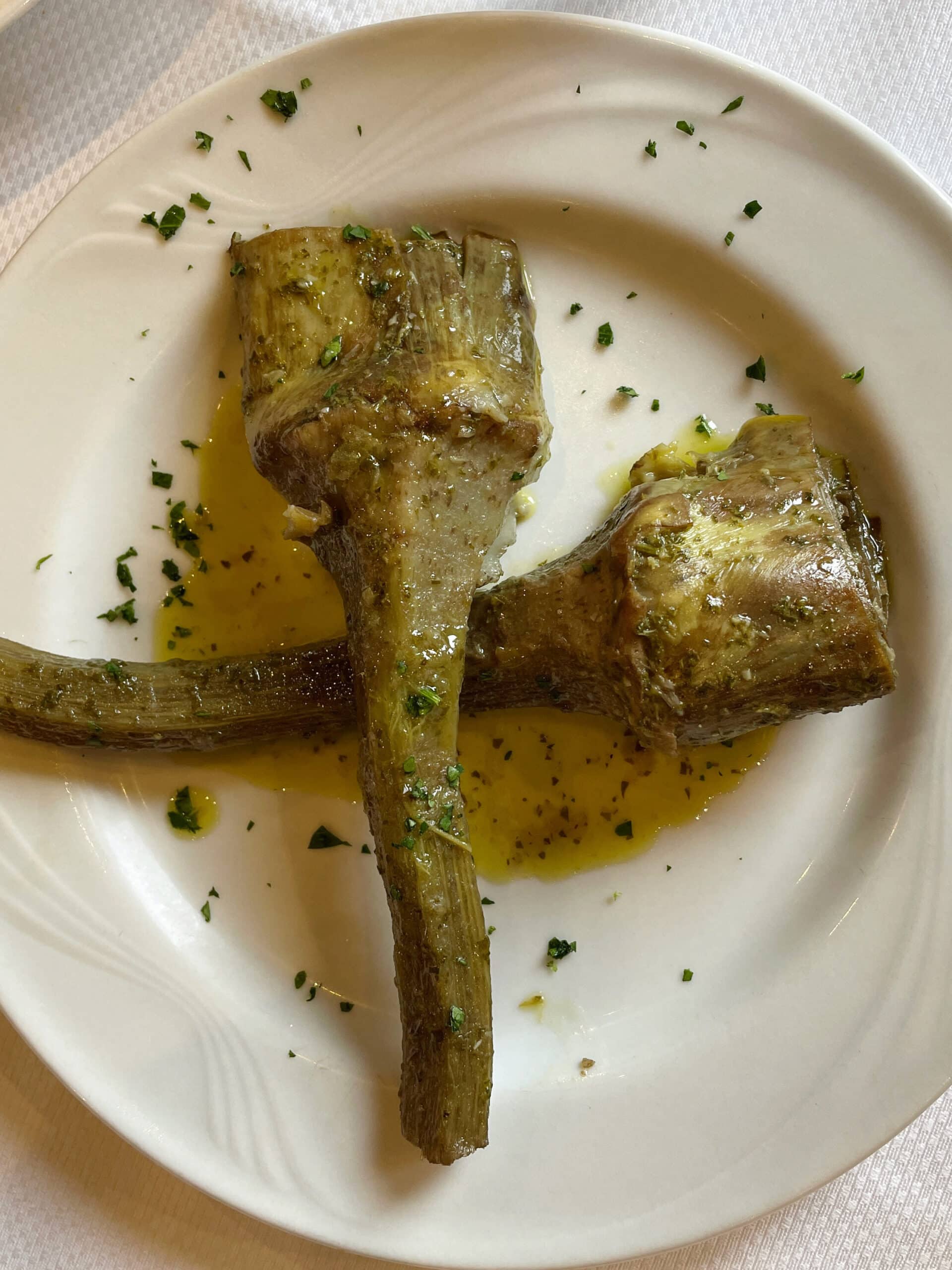 Two cooked artichoke stems with hearts, garnished with chopped herbs and served with olive oil, presented on a white plate.