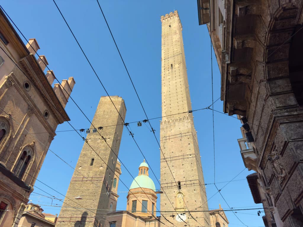 Two tall medieval brick towers, Le Due Torri, Torre Garisenda and Torre degli Asinelli, and a green-domed building rise against a clear blue sky, viewed from below with surrounding historic buildings and intersecting overhead wires. Visiting these towers is one of the best things to do in Bologna, Italy.