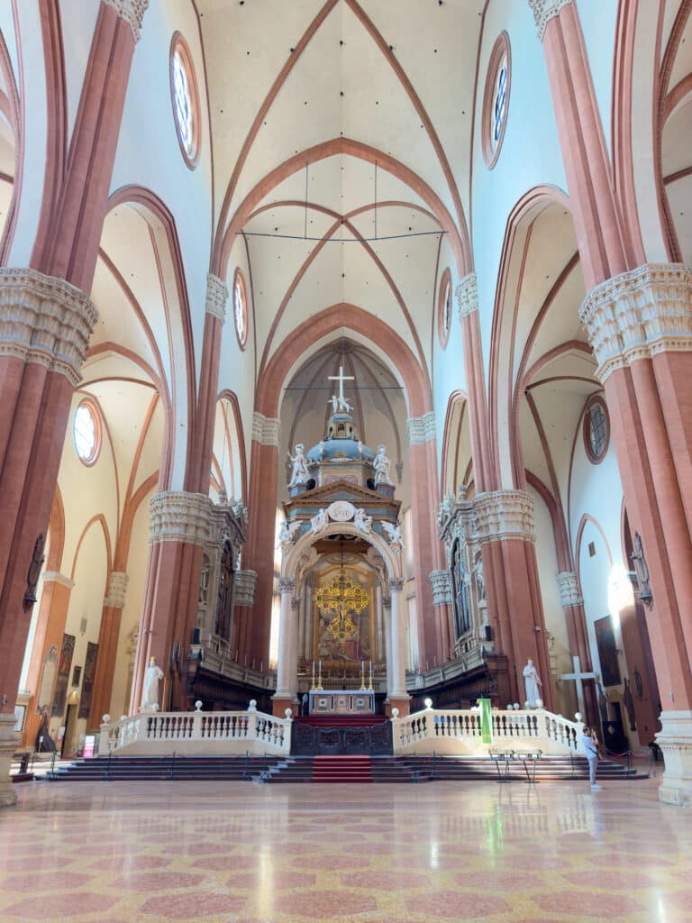 Grand interior of a cathedral with high vaulted ceilings, tall columns, arched windows, ornate altar, religious statues, and decorative floor patterns in warm tones.
