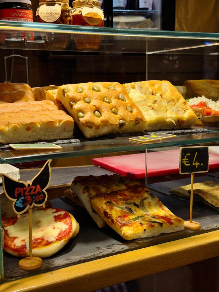 A bakery display case in Mercato di Mezzo with various types of focaccia bread, some topped with olives or onions, and pizza slices labeled with prices on small chalkboard signs.