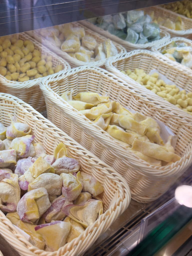 Several wicker baskets filled with various types of uncooked, colorful fresh pasta are displayed in a refrigerated glass case at a the Mercato di Mezzo in Bologna - one of the must-visits in Bologna, Italy.