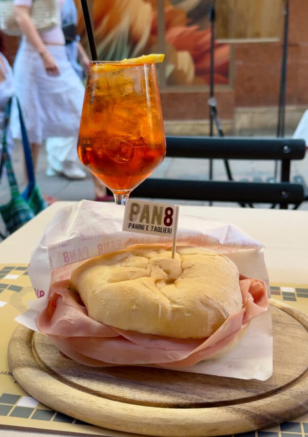 A round sandwich filled with sliced ham on a wooden plate, labeled "Pan8 Panini e Taglieri," sits beside an orange cocktail with ice and a straw on an outdoor café table. People walk by in the blurred background.