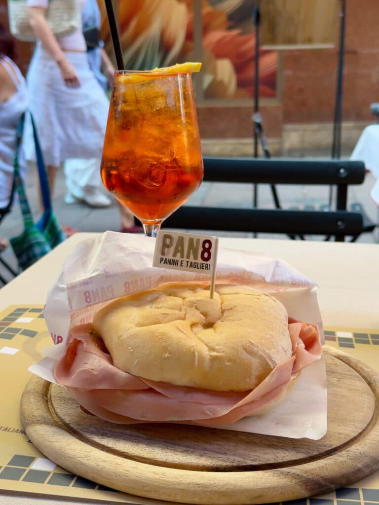 A round sandwich filled with sliced ham on a wooden plate, labeled "Pan8 Panini e Taglieri," sits beside an orange cocktail with ice and a straw on an outdoor café table. People walk by in the blurred background.