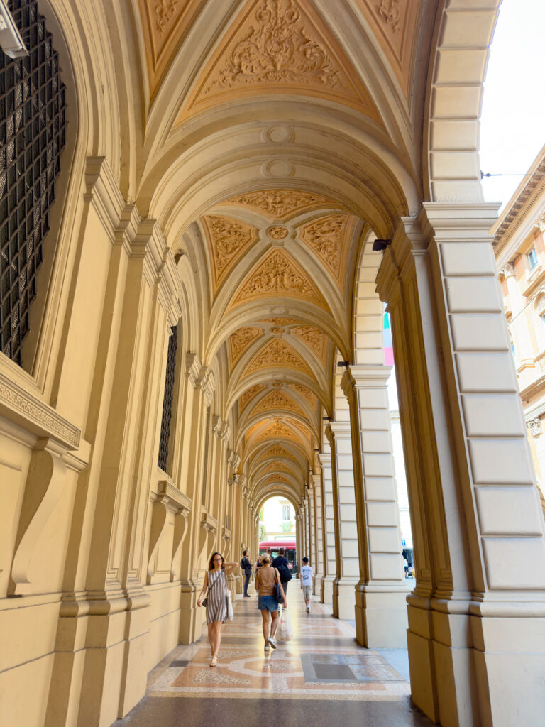 People walk under a grand covered walkway with ornate vaulted ceilings and porticoes of Bologna, lined with columns and detailed architectural designs, in a sunlit urban area.