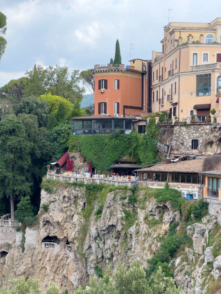 A restaurant in Tivoli, Ristorante il Ciocco, terrace with tables and red umbrellas is perched on a rocky cliff, surrounded by lush greenery and historic buildings in warm tones, overlooking a scenic landscape, Villa Gregoriana.