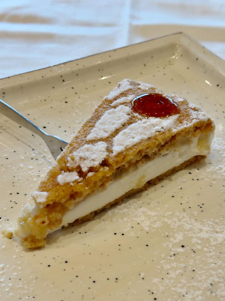 A slice of gluten free hazelnut cake with a creamy white ricotta filling, dusted with powdered sugar and topped with a small dollop of red jam, sits on a speckled plate with a fork.