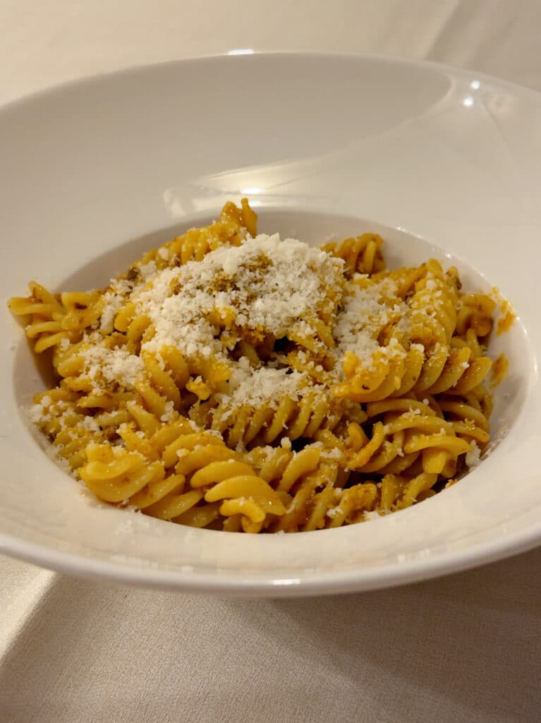 A white bowl filled with rotini pasta coated in a light sauce, topped with a generous sprinkle of grated cheese, sits on a light-colored tablecloth.