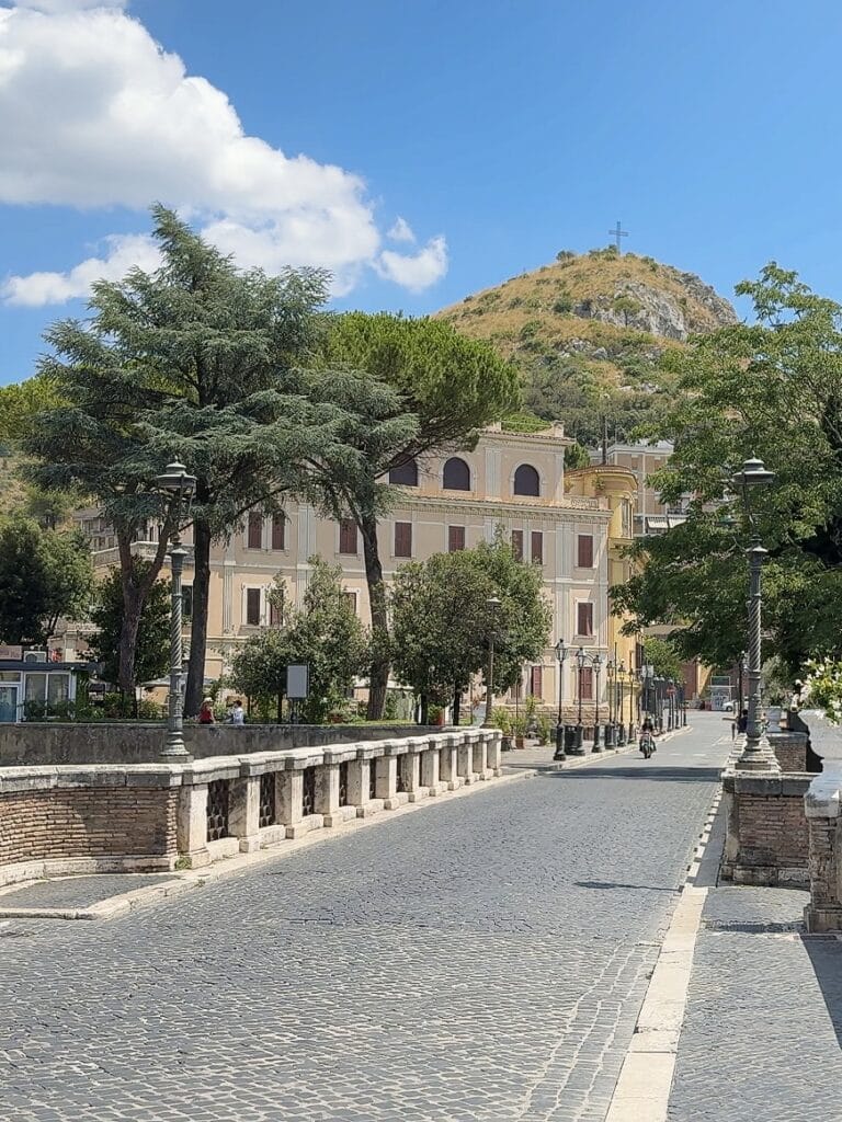 A cobblestone street in Tivoli, Italy with a stone railing runs toward pastel buildings and a tree-covered hill topped with a cross, under a blue sky with scattered clouds.