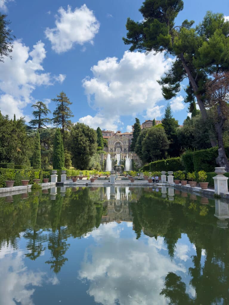 An ornate villa with fountains and lush gardens is reflected in a large rectangular pond, set beneath a bright blue sky with scattered clouds and surrounded by tall trees.
