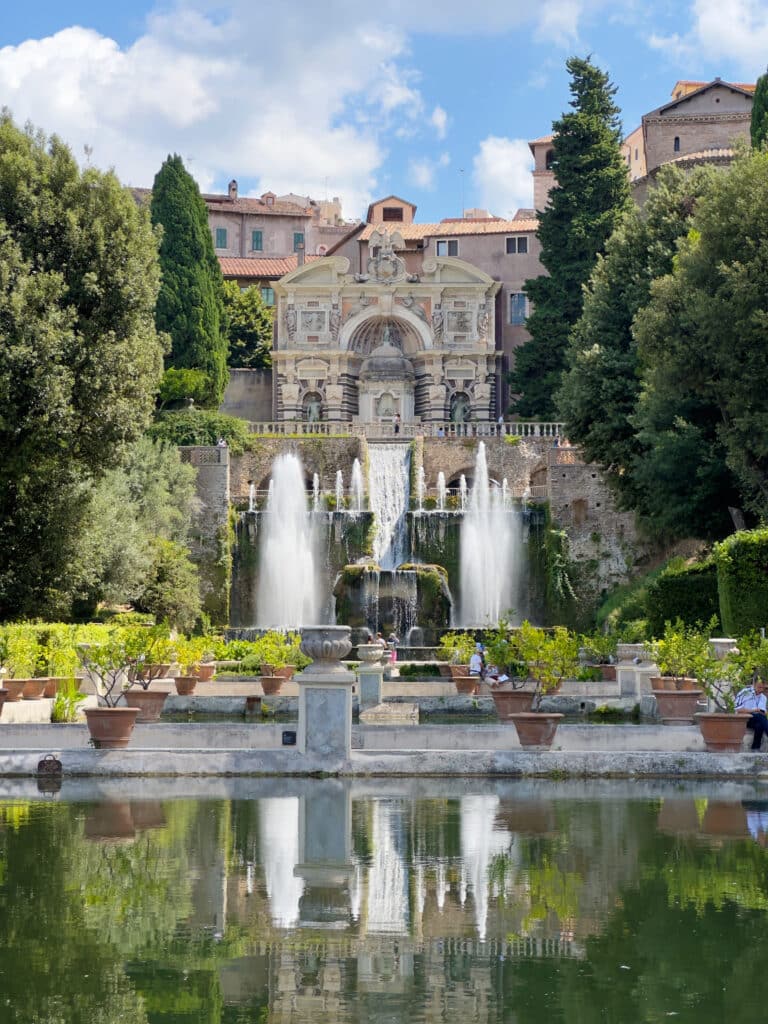 A grand stone fountain, Fontana di Nettuno and Fontana dell'Organo, with multiple water jets stands before a large historic villa, surrounded by lush greenery and reflected in a tranquil pond under a partly cloudy sky. This is a must visit on a Tivoli day trip.