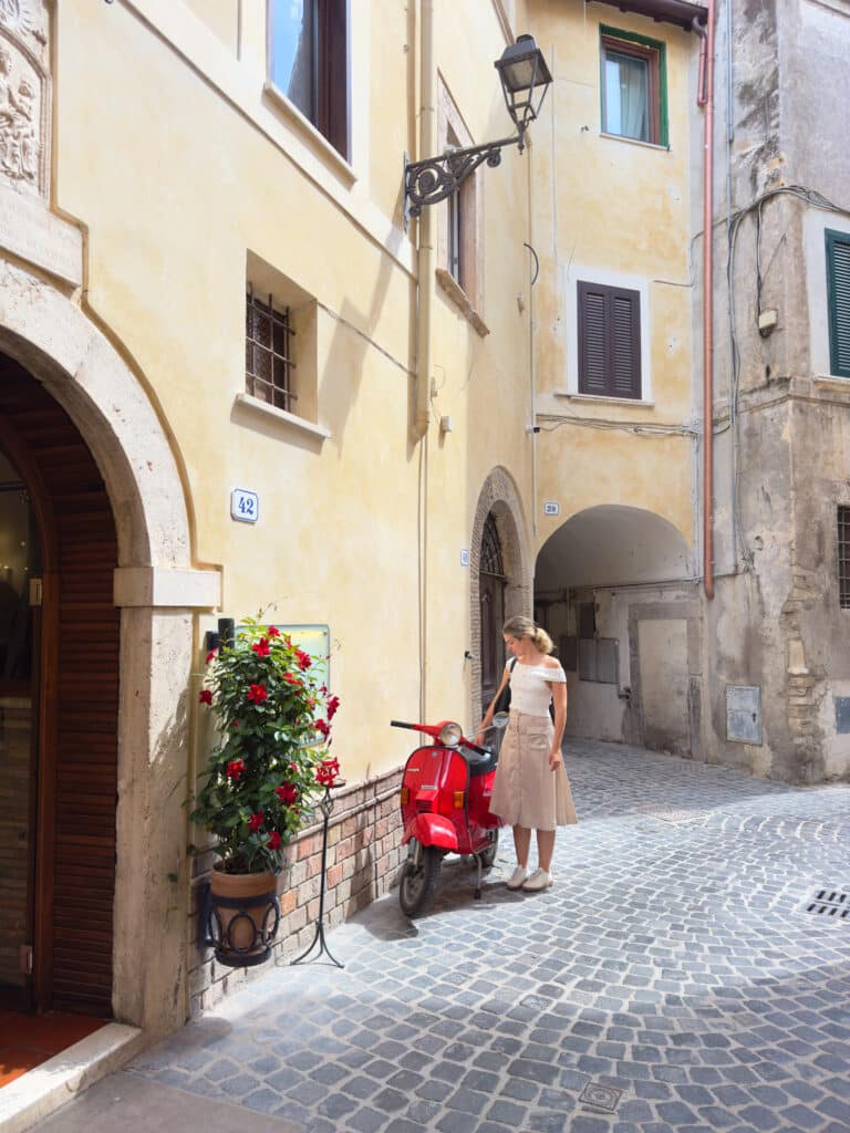 A woman in a light dress stands beside a red vespa scooter near a flower arrangement on a cobblestone street, surrounded by old yellow buildings with shuttered windows and arched doorways.