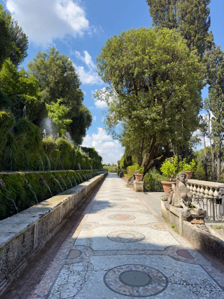 A sunlit stone pathway, Viale delle Cento Fontane in Villa d'Este of Tivoli, lined with trees and greenery, featuring a decorative mosaic floor, water fountains on the left, and stone statues and potted plants on the right under a blue sky with clouds. Tivoli is one of the best day trips from Rome.