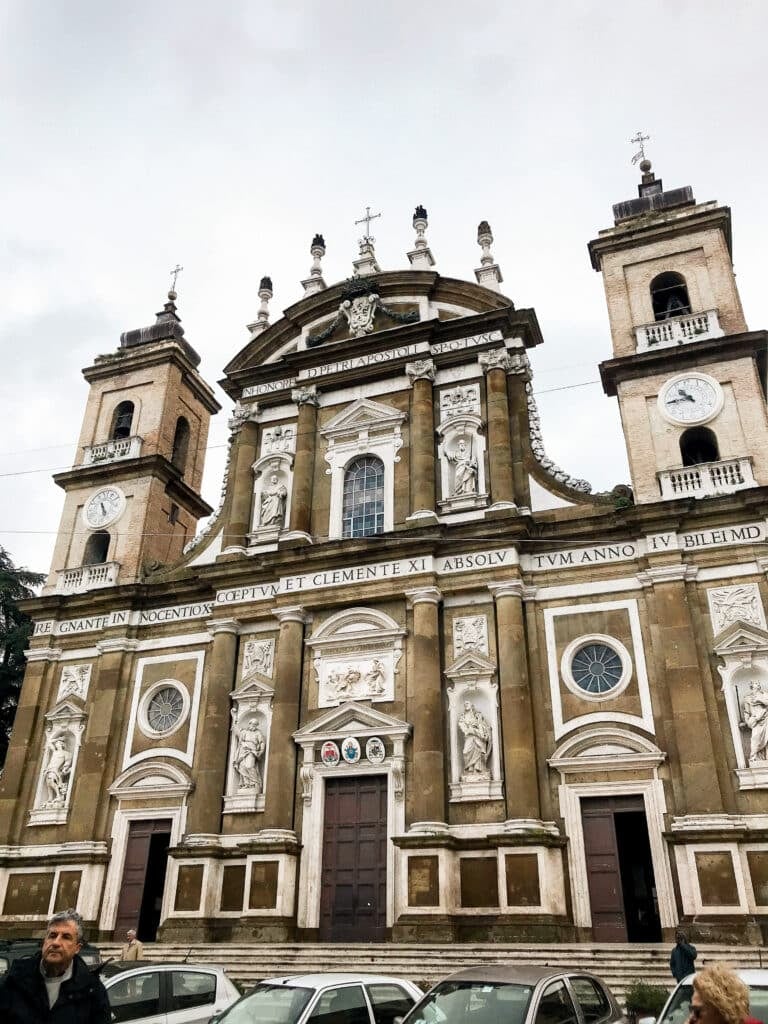 A historic church with two bell towers, ornate columns, statues, and three large wooden doors stands against an overcast sky. Several cars are parked in front, and a man is seen walking by.