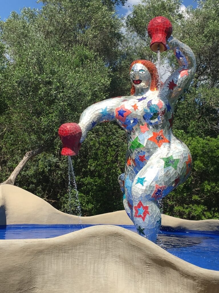 A colorful mosaic sculpture in the Giardino dei Tarocchi (Tarot Gardens) of a woman holding two red jugs, with water pouring from them, stands in a blue pool surrounded by greenery and beige curved shapes. The figure is decorated with stars and vibrant patterns. The Giardino dei Tarocchi is one of the best day trips from Rome, Italy by car.