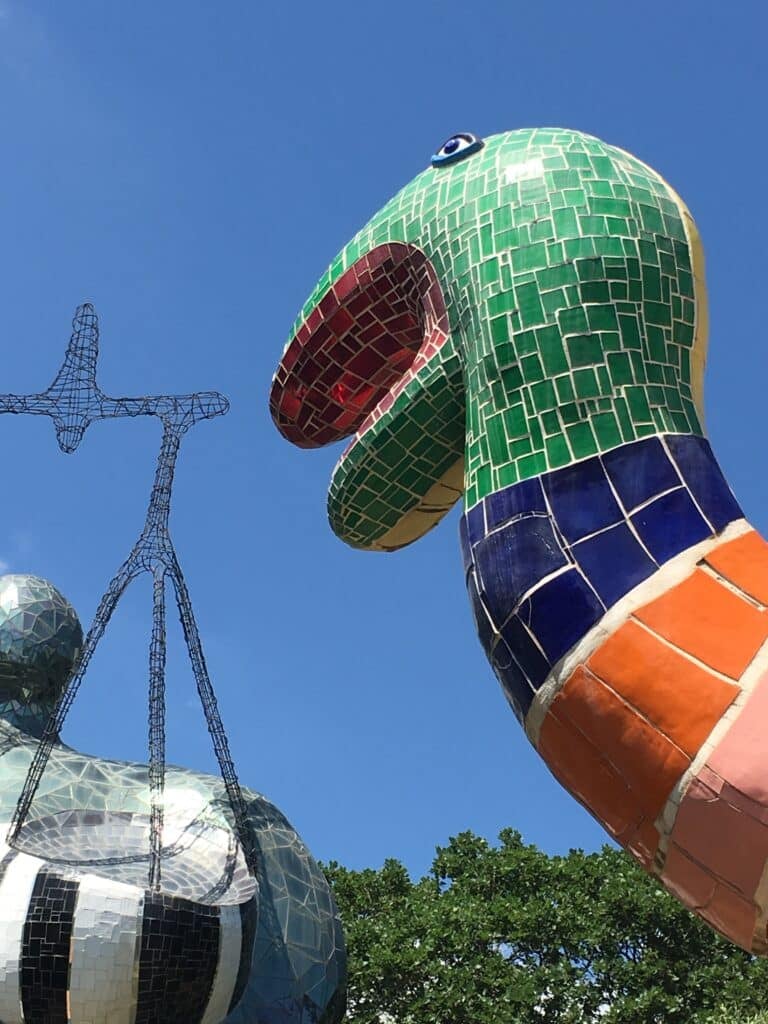 Mosaic-tiled sculpture of a large, colorful snake-like creature with an open mouth, in the Giardino dei Tarocchi (Tarot Garden) facing a wireframe human figure holding a telescope, set against a clear blue sky and green trees.