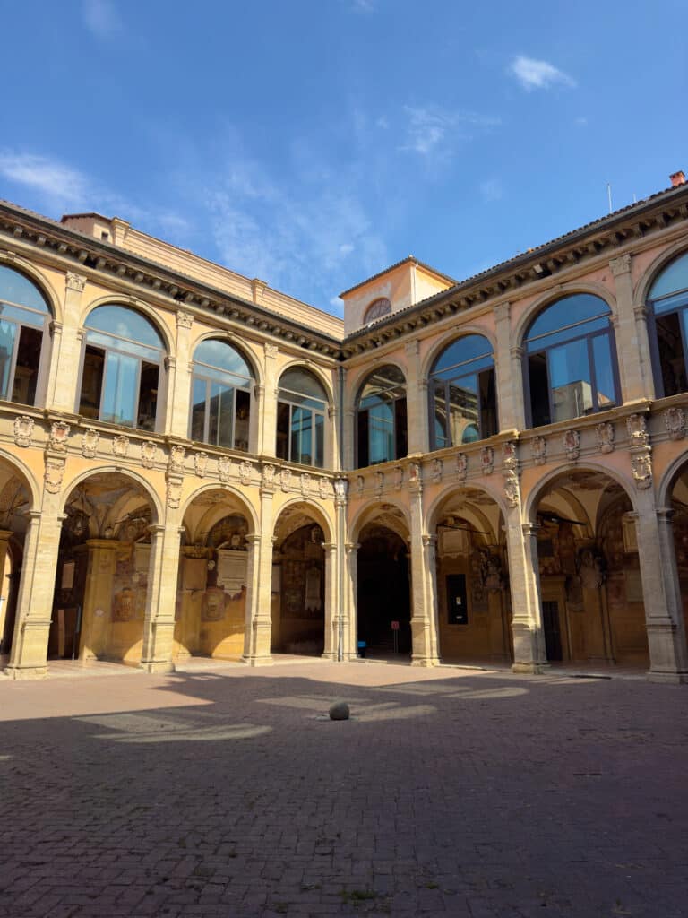 Sunlit courtyard of a historic building, Palazzo Archiginnasio, with arched columns and upper-level windows. The walls feature decorative details, and the sky is clear and blue above the structure. This is a must visit when spending one day in Bologna.