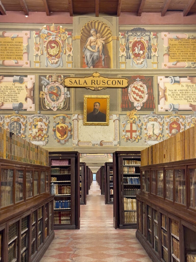 Ornate library hall with wooden bookshelves full of books, decorative painted crests and murals on the upper walls, and a portrait above a sign reading “Sala Rusconi.” Marble floor and vaulted ceiling visible. This is the oldest library of Bologna and is within the Palazzo Archiginnasio.