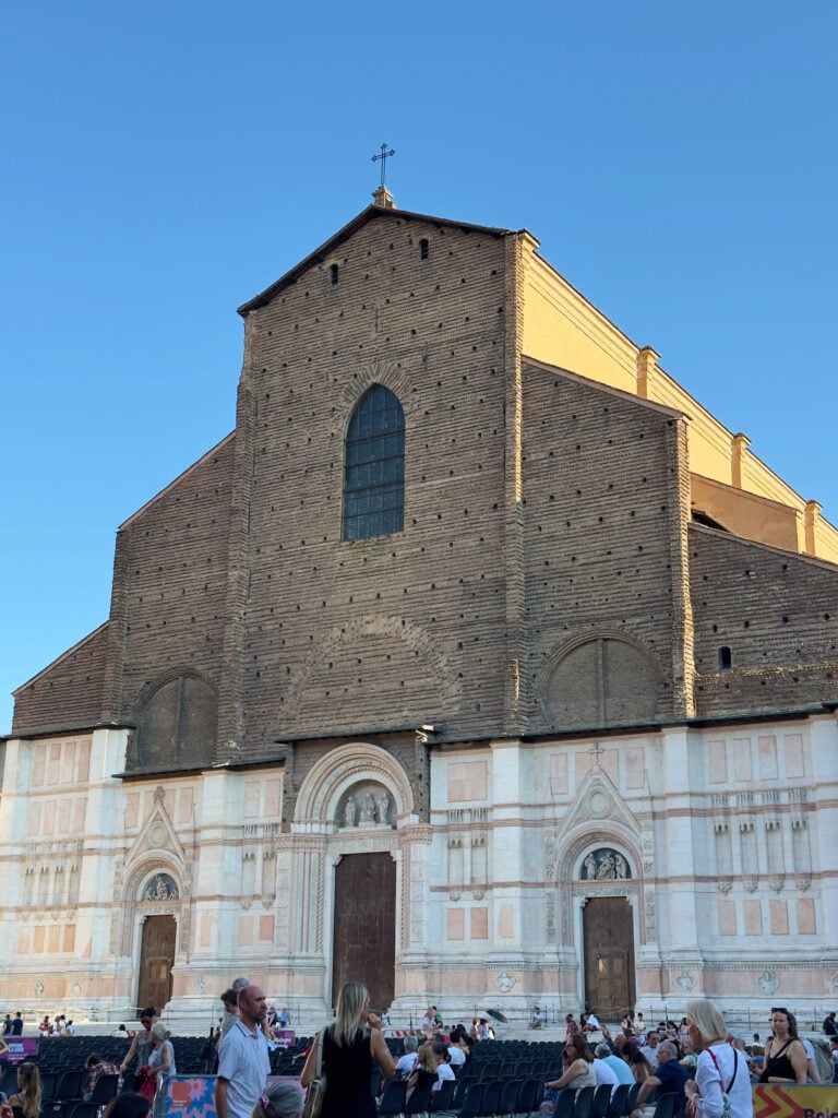 A large brick basilica, Basilica di San Petronio in Bologna, with a partly unfinished façade stands against a clear blue sky. People gather in the open square in front, some sitting and others standing, enjoying the outdoor setting. This is a must visit when spending one day in Bologna.