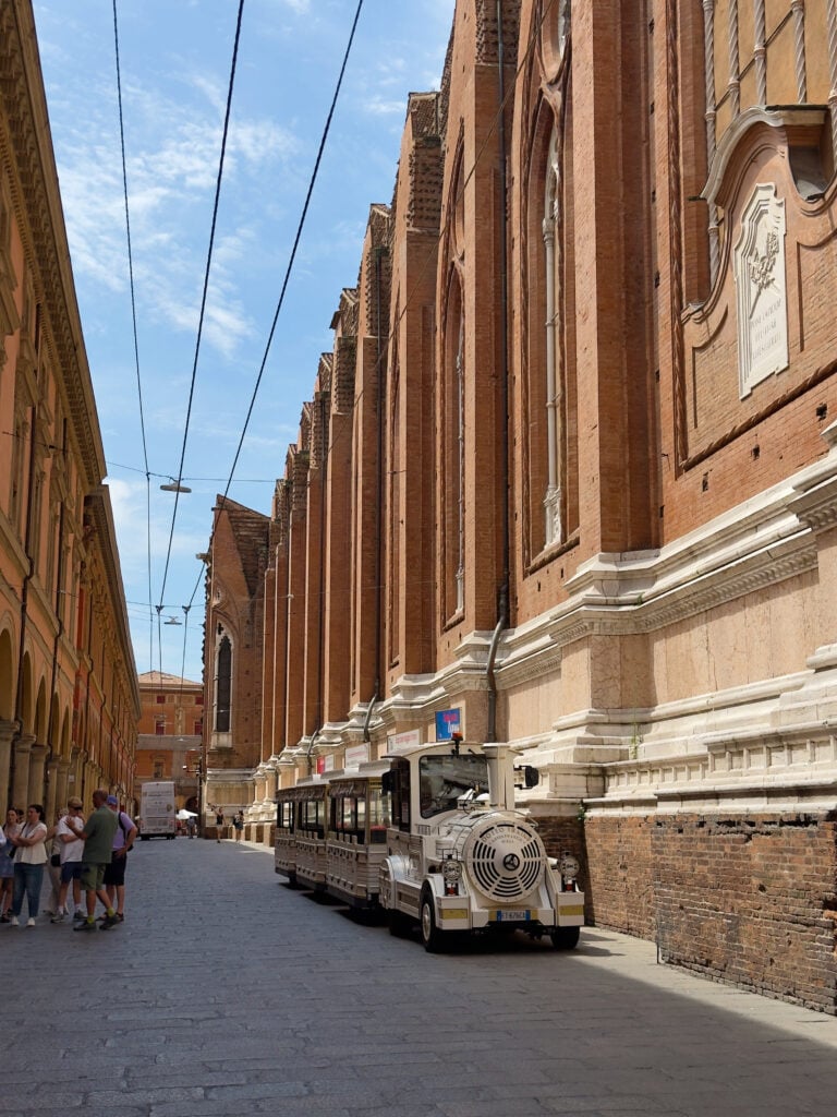 A small tourist train is parked on a narrow cobblestone street in Bologna lined with tall historic brick buildings under a blue sky. A group of people stands on the left side of the street.