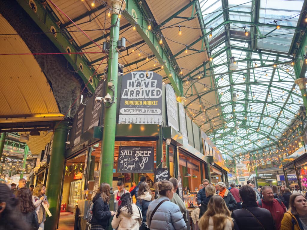 Indoor view of Borough Market, London, with crowds of people, green metal beams, food stalls, and a large sign that reads “YOU HAVE ARRIVED BOROUGH MARKET NORTHFIELD FRIDA”—a must-see in London for first time visitors 