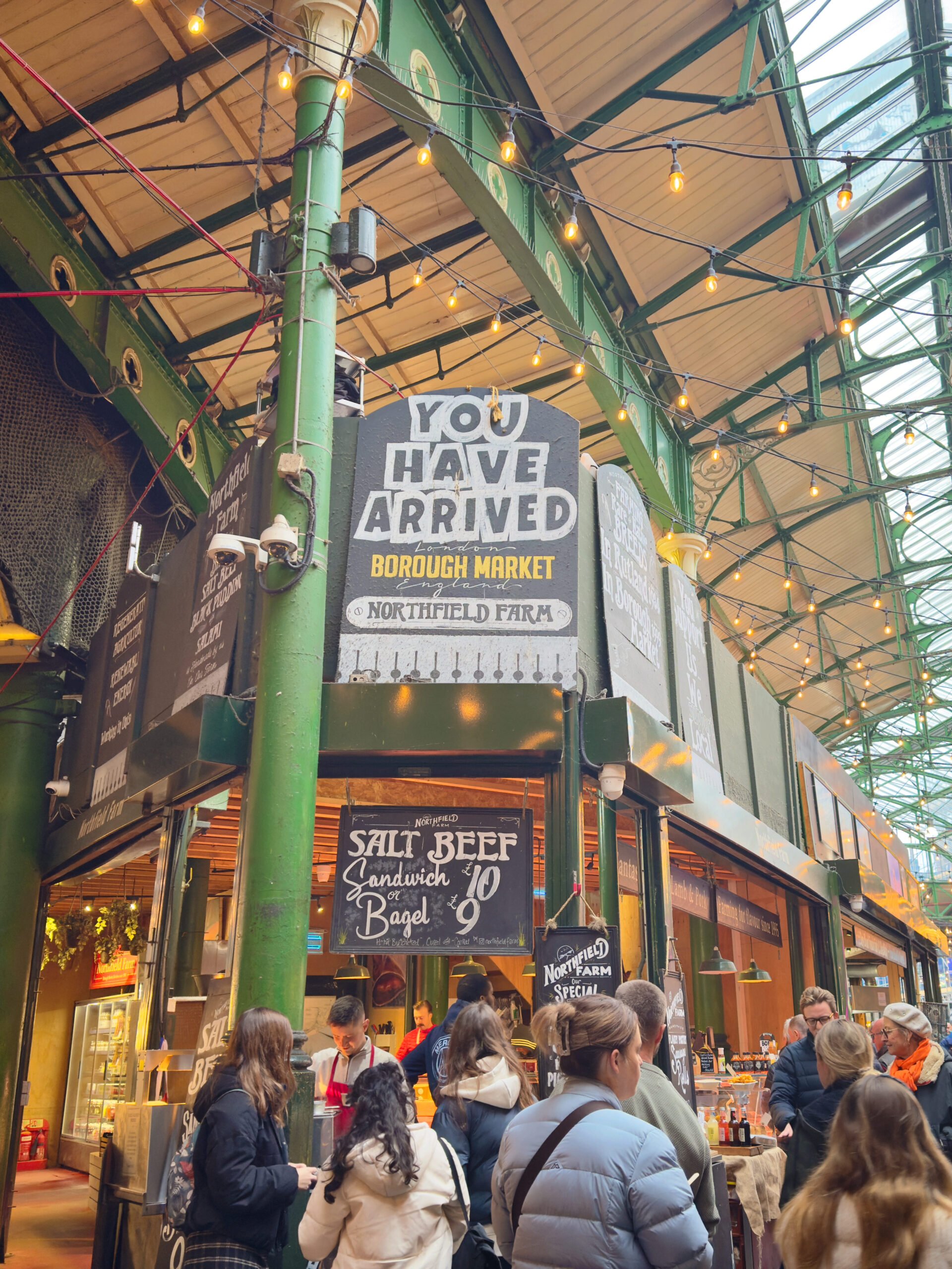 Indoor market scene at Borough Market with a large "You Have Arrived" sign overhead, several people queuing at a food stall beneath string lights, and colorful green metal columns supporting the roof. This is one of the best things to do in Southwark