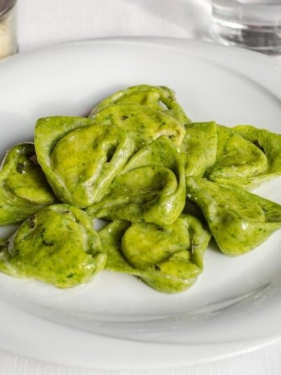 A plate of green, heart-shaped ravioli with a light sauce sits on a white tablecloth, accompanied by a glass of water, a glass of red wine, and a container of grated cheese.