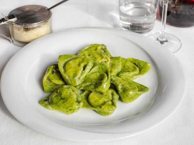 A plate of green, heart-shaped ravioli with a light sauce sits on a white tablecloth, accompanied by a glass of water, a glass of red wine, and a container of grated cheese.