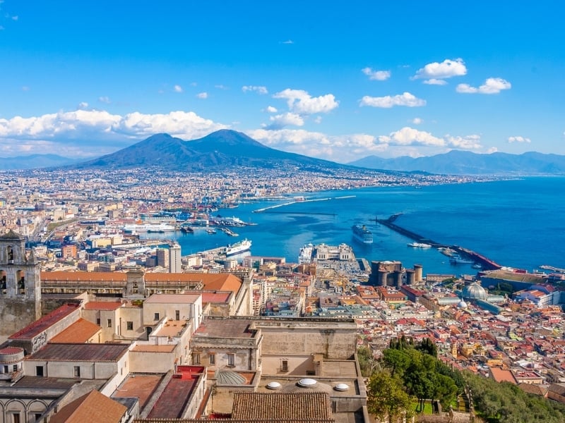 A panoramic view of Naples, Italy with colorful buildings, a busy port, and the blue waters of the bay. Mount Vesuvius rises in the background under a bright, partly cloudy sky.