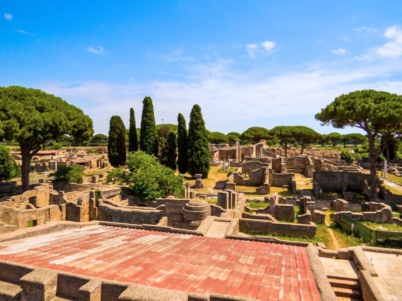 Ruins of ancient stone buildings surrounded by tall cypress and pine trees under a bright blue sky, showing an archaeological site with scattered columns and walls.