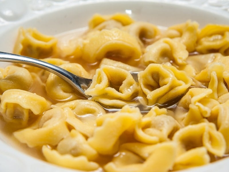 A close-up of a bowl of tortellini in Brodo, a classic Bologna food, with yellow pasta in broth and a spoon lifting several pieces. The tortellini have a folded, ring-like shape and are floating in the light-colored liquid.