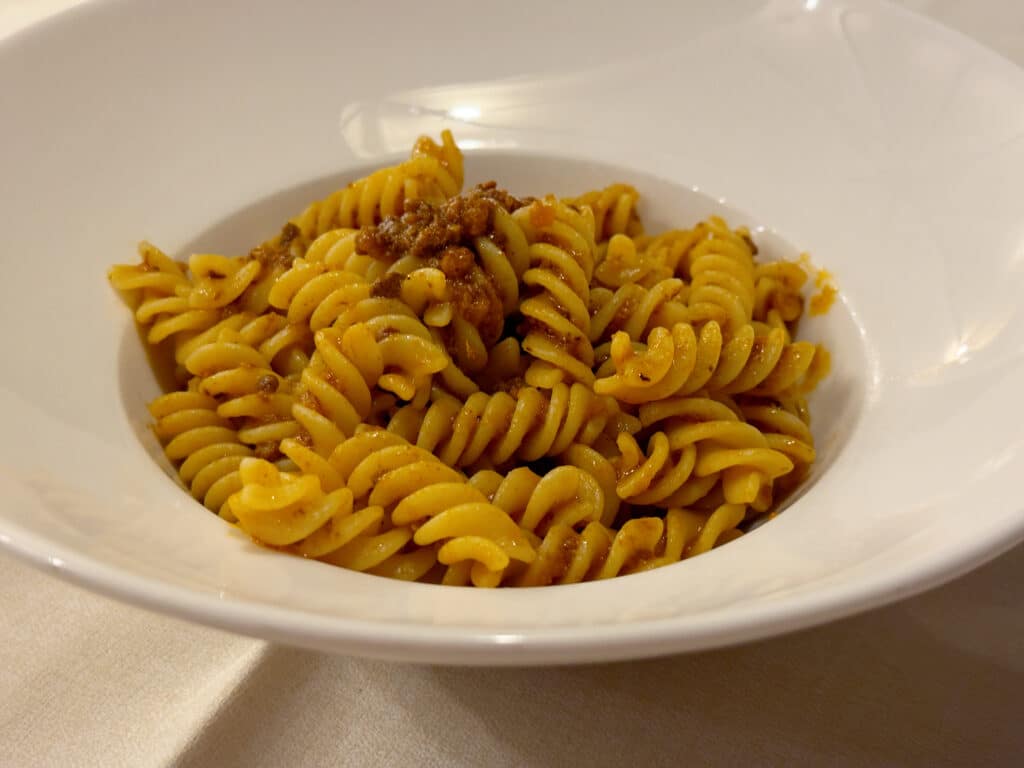 A white bowl filled with cooked fusilli pasta topped with a brown meat sauce, placed on a light-colored tablecloth.