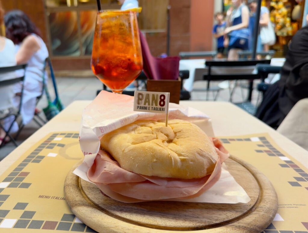 A sandwich filled with sliced deli meat sits on a wooden plate with a “PAN8” flag. An orange drink with ice is in the background, and people are walking past outdoor café tables on a city street.