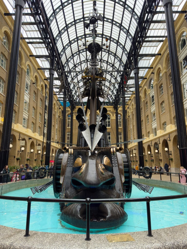 A large mechanical sculpture featuring gears and clock parts stands in a circular pool under a glass roof, surrounded by tall brick buildings and people observing. Visiting Hay's Galleria is one of the best things to do in Southwark