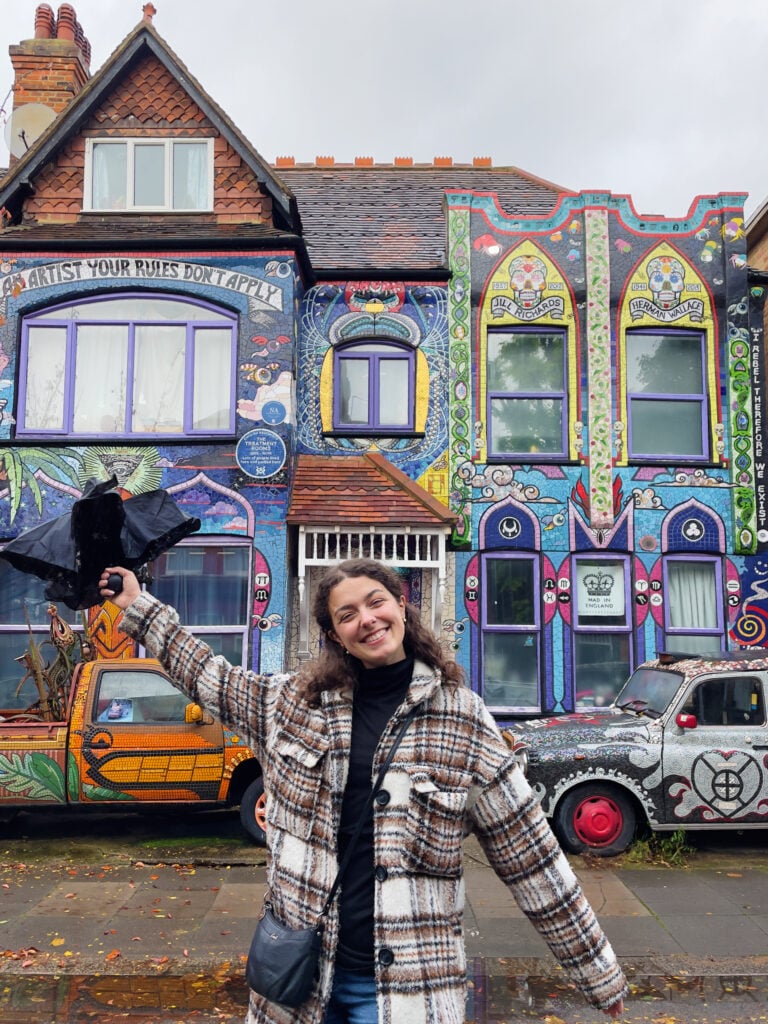 A person in a plaid coat smiles and poses with an arm raised, holding a black bird prop, in front of a colorful, mural-covered house and a painted car on a cloudy day. Showing what she wears in London, lots of layers.