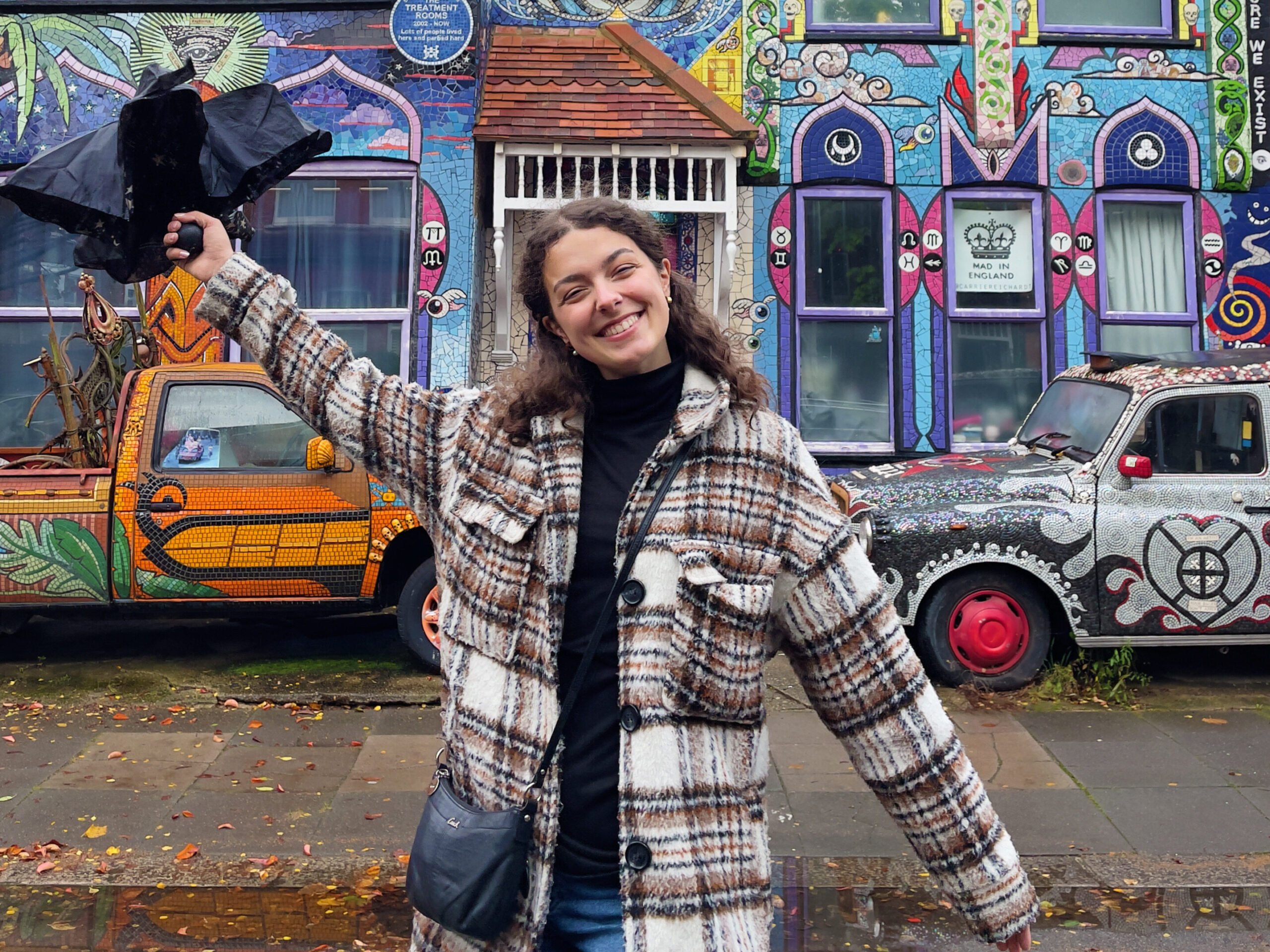 A person in a plaid coat smiles and poses with an arm raised, holding a black bird prop, in front of a colorful, mural-covered house and a painted car on a cloudy day.