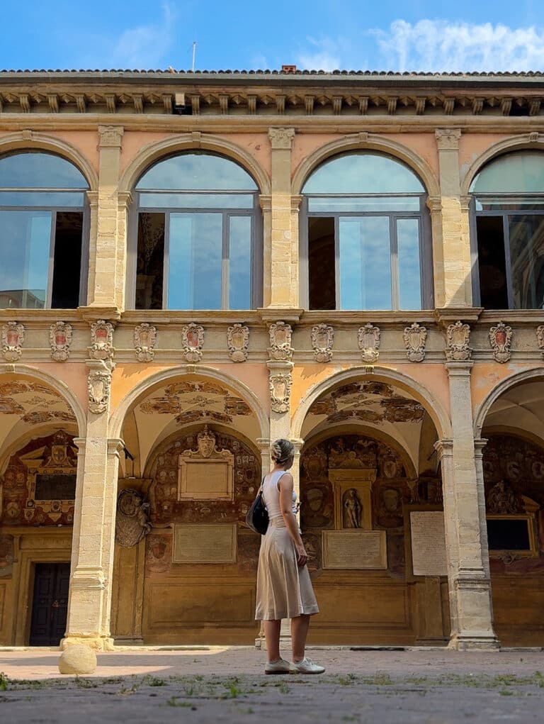 A woman in a light dress stands in a courtyard, looking up at an old building with arches, columns, and decorated walls, under a clear blue sky.