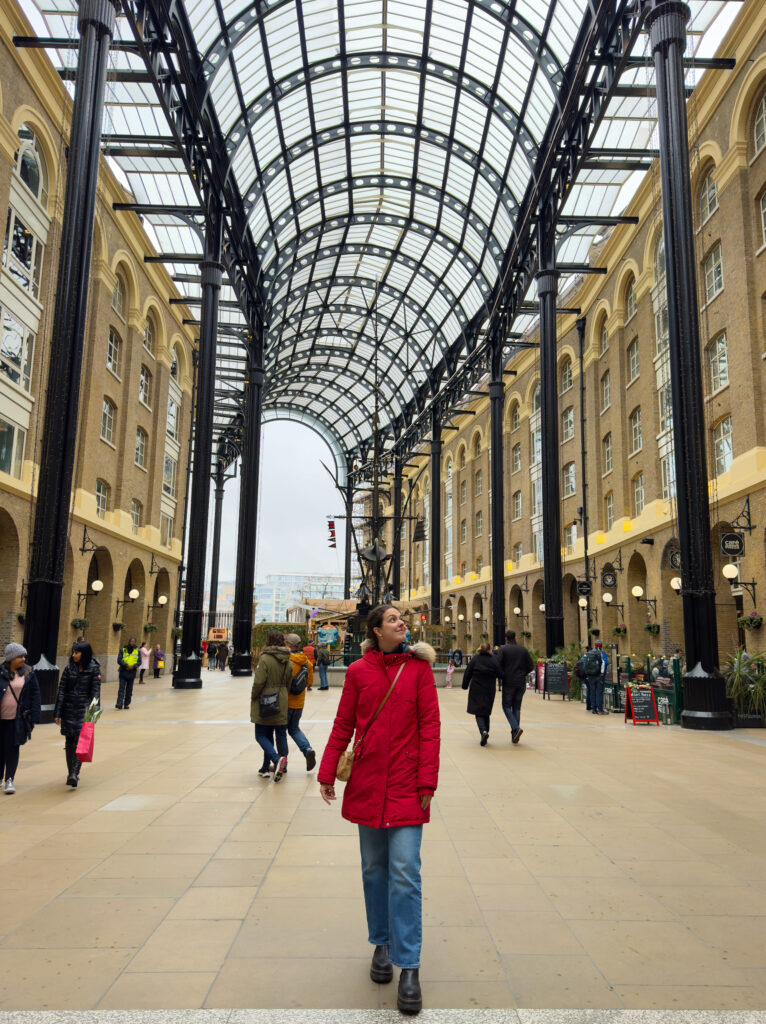 A person in a red coat walks under a large glass and steel arched roof between tall brick buildings, with other people walking and shops visible in the background. Visiting Hay's Galleria is one of the best things to do in Southwark