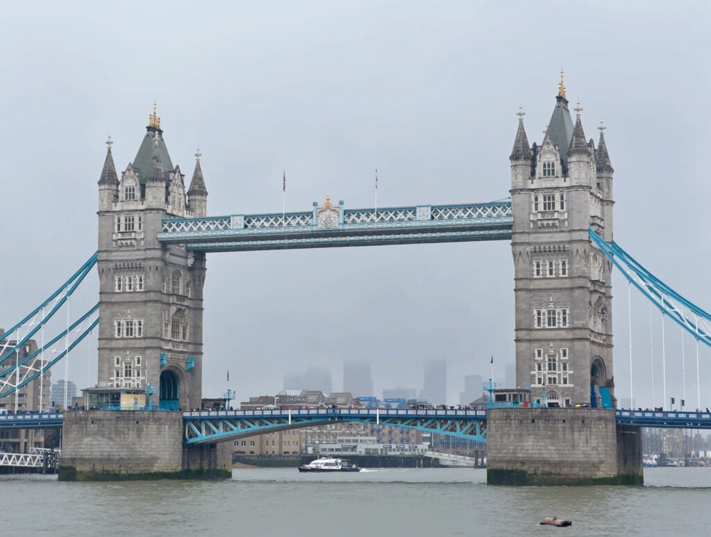 Tower Bridge in London on a cloudy day, with two tall towers connected by a blue walkway and suspension elements. The River Thames flows below, and a boat is seen passing under the bridge. City buildings are faint in the background. This London icon is one of the best attractions to visit in Southwark borough.