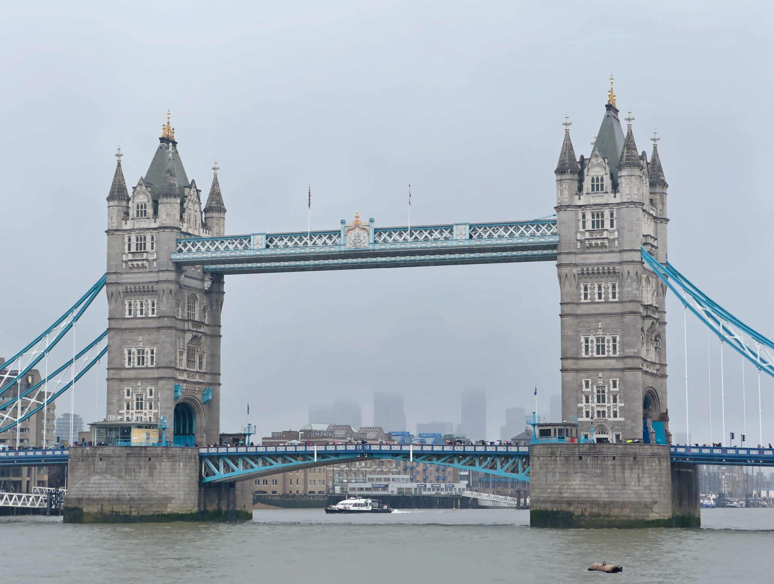 Tower Bridge in London on a cloudy day, with two tall towers connected by a blue walkway and suspension elements. The River Thames flows below, and a boat is seen passing under the bridge. City buildings are faint in the background.