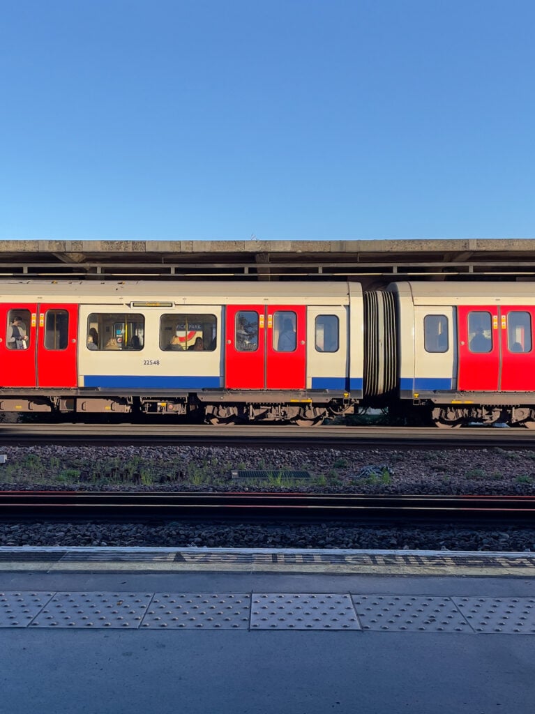 A red, white, and blue train stopped at an outdoor station under a clear blue sky, with empty tracks and the platform in the foreground.