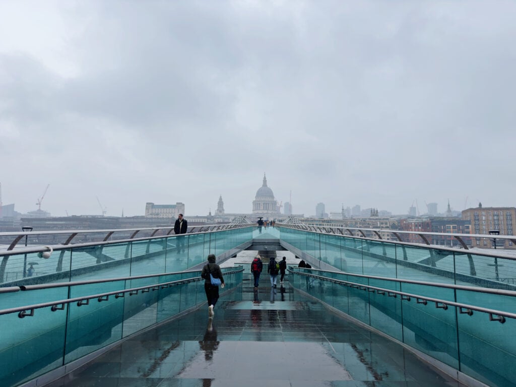 People walk across the wet Millennium Bridge in London on a cloudy day, with St. Paul’s Cathedral visible in the background through the mist.