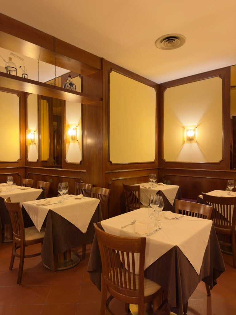A cozy restaurant dining area with wooden chairs and tables covered in white and brown tablecloths. Wine glasses and napkins are set on each table. Warm wall lights and wooden paneling create an inviting atmosphere.