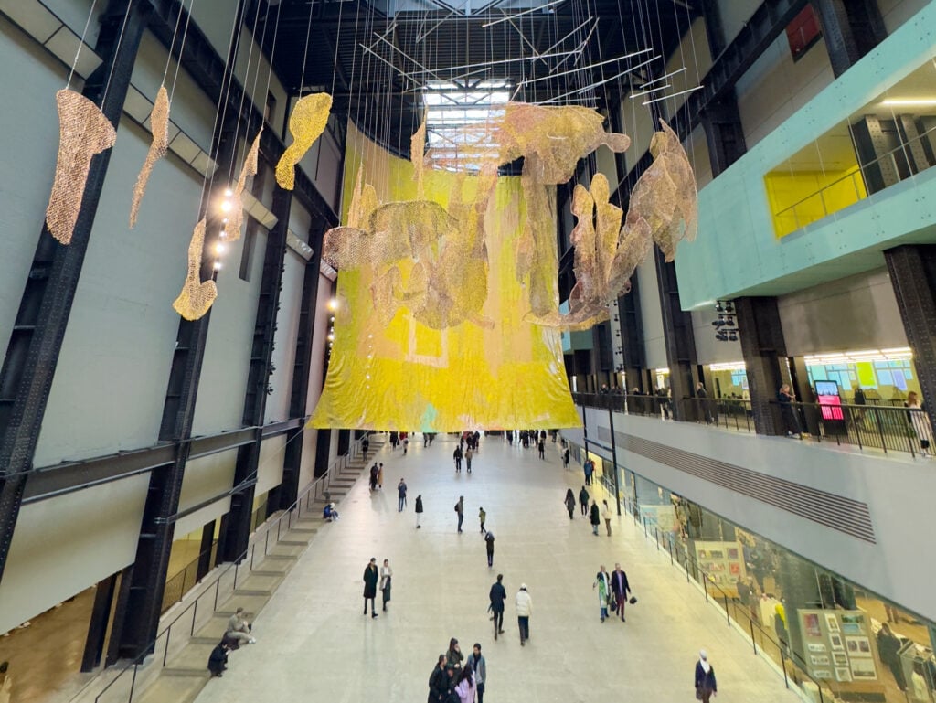 Large yellow and gold art installation hangs in an industrial-style atrium with high ceilings and tall windows as people walk and gather below on the concrete floor of Tate Modern museum in Southwark, London.