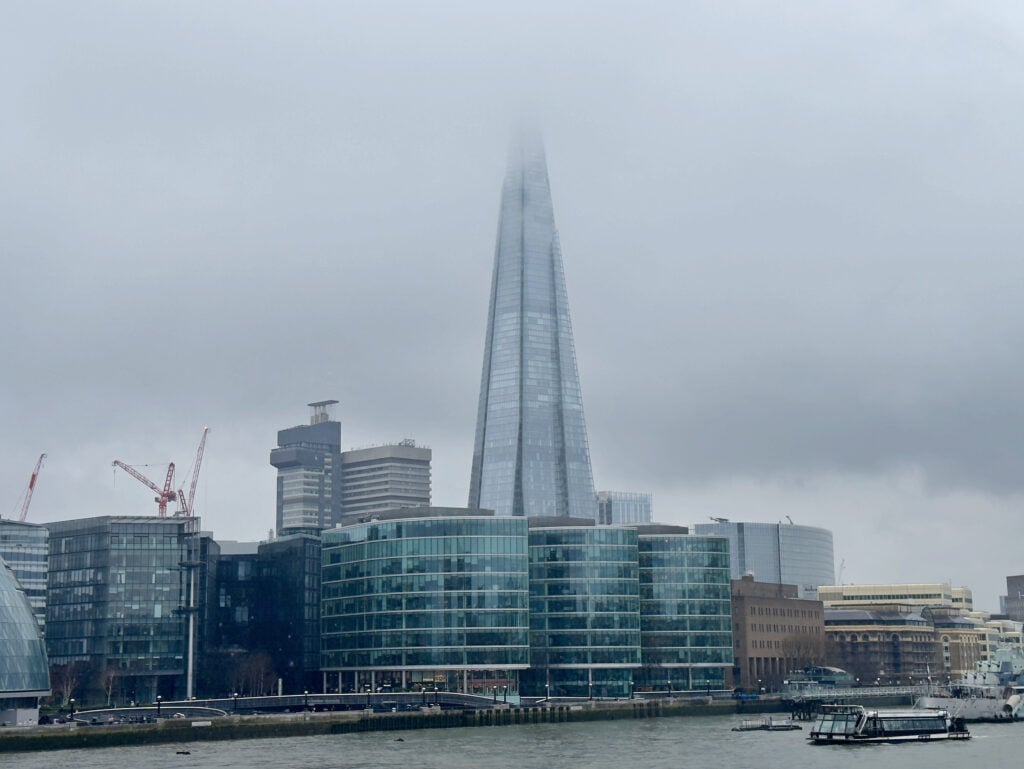 The Shard skyscraper in London rises above nearby glass buildings, its top obscured by thick fog on a grey, overcast day. Cranes and the River Thames are visible in the foreground. This is one of the best things to do in Southwark