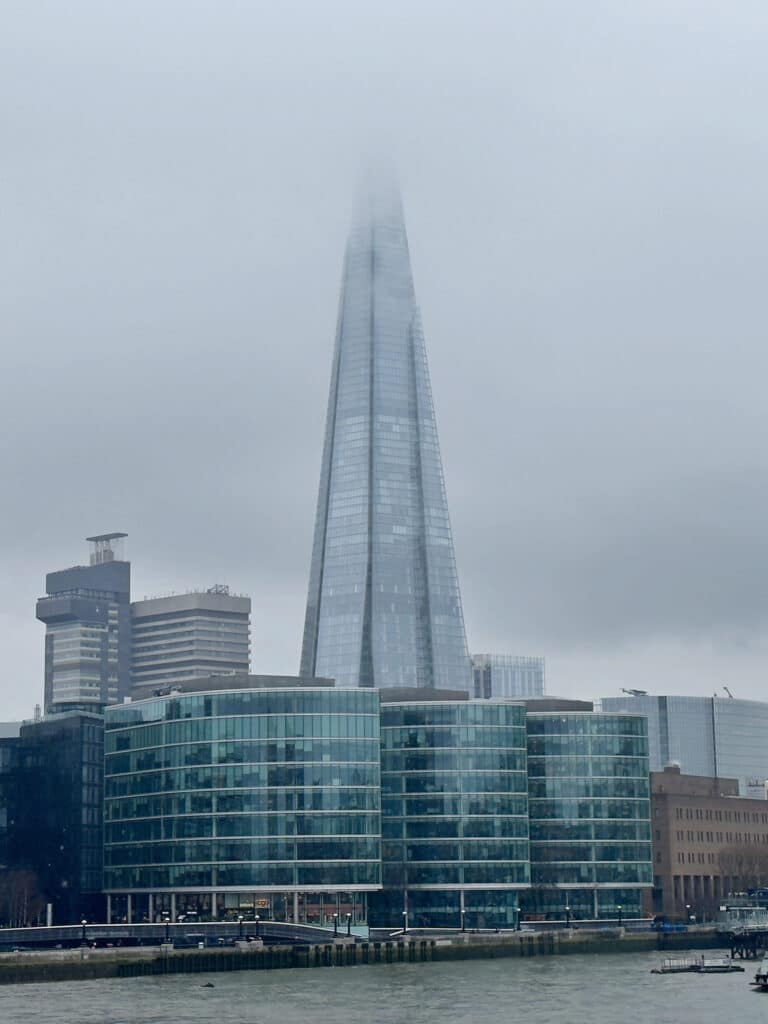 The Shard skyscraper in London rises above nearby glass buildings, its top obscured by thick fog on a grey, overcast day. Cranes and the River Thames are visible in the foreground.