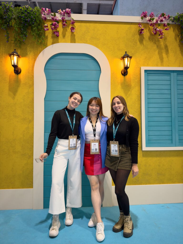 Three young women, travel bloggers, with name badges pose and smile in front of a bright yellow wall decorated with an arched door, blue shutter, two lanterns, and hanging pink flowers. They are attending World Travel Market (WTM) London.