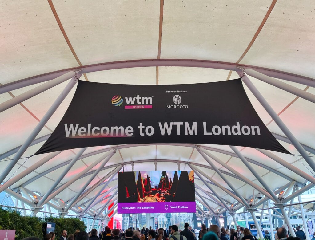 A large black banner reading "Welcome to WTM London" hangs under a modern, white canopy at an event entrance. People gather below, and a screen displays event information for World Travel Market London 2025 in the background.